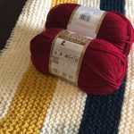 handknit hudson's bay blanket with 2 balls of red yarn sitting on top